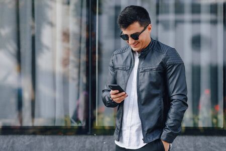 young stylish guy in glasses in black leather jacket with phone on simple glass backgroundの写真素材