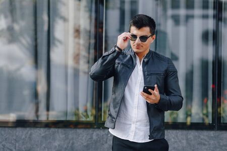 young stylish guy in glasses in black leather jacket with phone on simple glass backgroundの写真素材