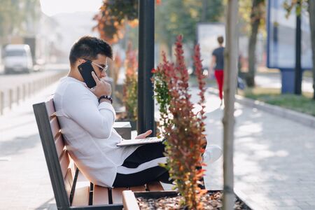 young stylish guy in white shirt with phone and notebook works on bench on sunny warm day outdoors, freelanceの写真素材