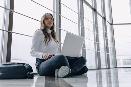 attractive girl working with laptop and things in airport terminal or office on floor. travel atmosphere or alternative work atmosphere. freelancer student travels to business meeting. concept of alternative workplace, technology and waiting.の写真素材