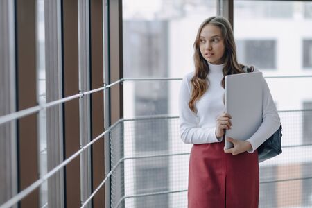 attractive girl walks in a bright corridor with laptop and things in airport terminal or office. travel atmosphere or alternative work atmosphere. freelancer student travels to business meeting with technology.の写真素材