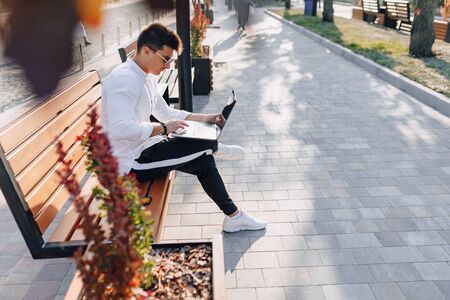 young stylish guy in white shirt with phone and notebook works on bench on sunny warm day outdoors, freelanceの写真素材