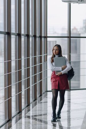 attractive girl walks in a bright corridor with laptop and things in airport terminal or office. travel atmosphere or alternative work atmosphere. freelancer student travels to business meeting with technology.の写真素材