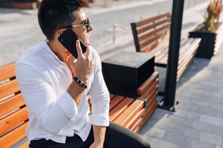 young stylish guy in white shirt with phone on bench on sunny warm day outdoorsの写真素材