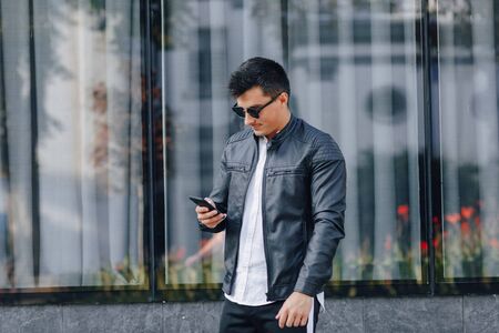 young stylish guy in glasses in black leather jacket with phone on simple glass backgroundの写真素材