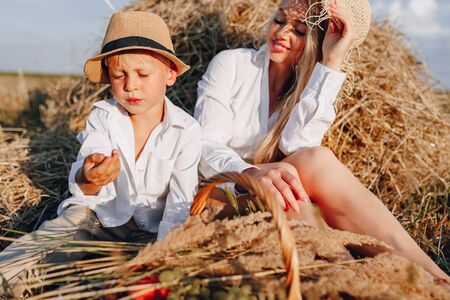 pretty blond long-haired woman with little blond son at sunset relaxing in the field and savoring fruit from a straw basket. summer, farming, nature and fresh air in the countryside. sunset.の写真素材
