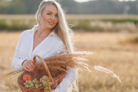 A pretty blond woman with long hair in a field at sunset carries a fruit basket and a bouquet of straw. summer, farming, nature and fresh air in the countryside. sunny.の写真素材