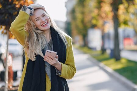 cute emotional attractive blonde woman in coat with smartphone walks the city street. communication during the walk, lifestyle, street in good weather.の写真素材