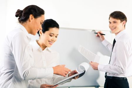 Two successful business ladies looking at notepad on the background of smiling man standing by whiteboard with paper in handの写真素材