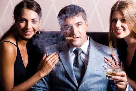Portrait of successful man smoking a cigar holding whisky with pretty women near by の写真素材