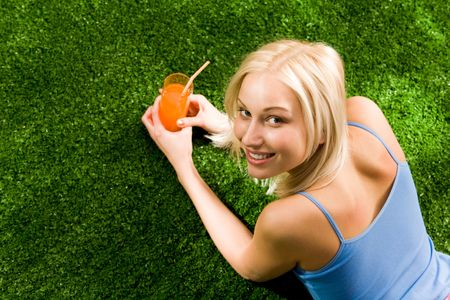 Photo of happy blond female lying on green grass with glass of juice and looking at cameraの写真素材