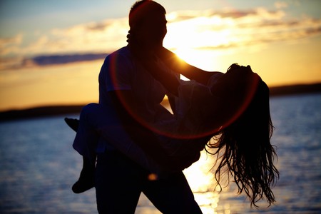 Silhouettes of happy guy holding his girlfriend by the lake at sunsetの写真素材