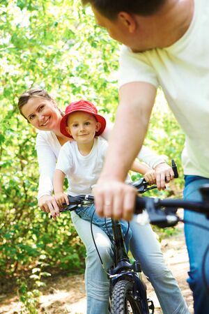 happy woman with son riding a bicycle in park while looking at her husbandの写真素材