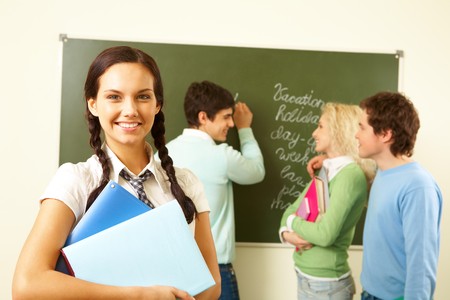 Portrait of cute girl with textbooks on background of communicating groupmates in classroomの写真素材