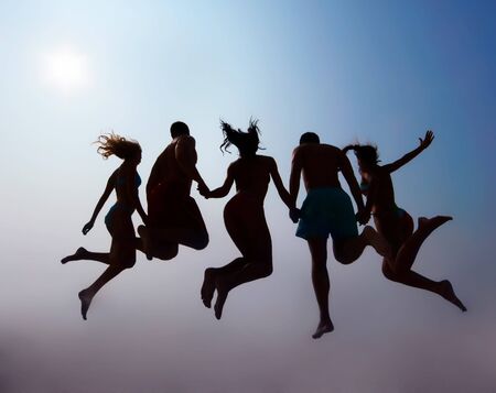 Rear view of friends holding by hands and jumping on sandy beach against sky with sun on itの写真素材