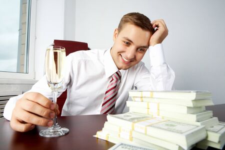 Image of young successful employer looking at heap of dollar bills and touching glass with champagne on workplaceの写真素材