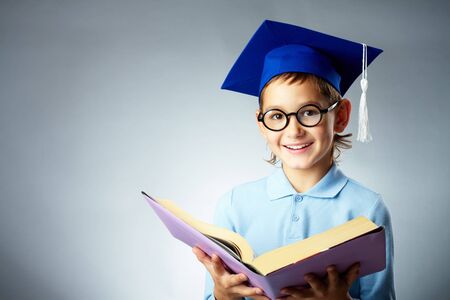 Portrait of cute lad in eyeglasses and student hat holding open book