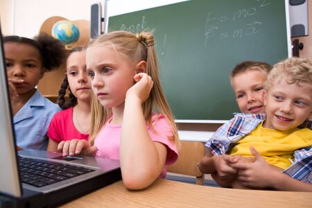 Photo of smart schoolgirl looking at the laptop with her classmates on background 