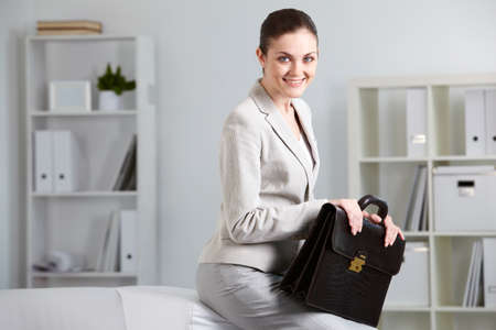 Portrait of smiling businesswoman with briefcase looking at camera in officeの写真素材