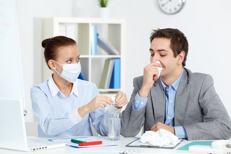 Image of sick businessman with tissue looking at his colleague in mask dissolving solution for him in officeの写真素材