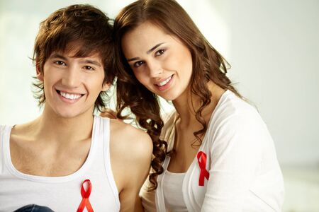 Smiling guy and girl with red ribbon badges sitting together and looking at camera