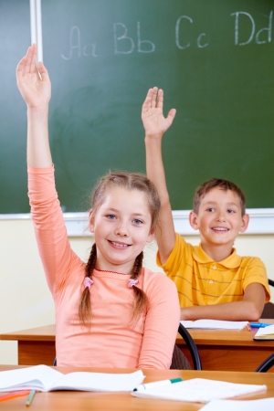 Portrait of lovely girl and her classmate on background raising hands at lessonの写真素材
