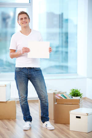 A young guy showing blank paper in new flat surrounded with boxes