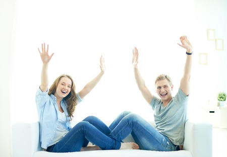 Joyful couple sitting on sofa with raised arms and looking at cameraの写真素材