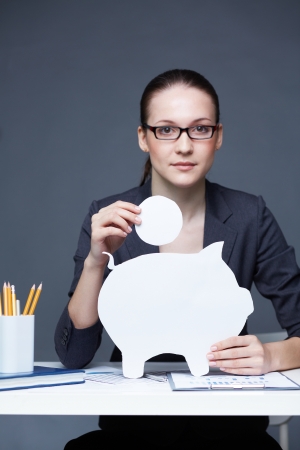 Portrait of young businesswoman with paper piggy bank and coin looking at cameraの写真素材