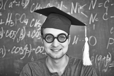 Black-and-white image of handsome student in graduation hat and eyeglasses looking at camera on background of chalkboardの写真素材