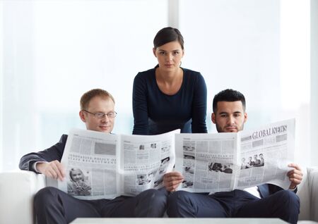 Two handsome businessmen reading newspapers in office with serious businesswoman looking at camera near byの写真素材