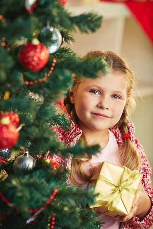 Adorable girl with giftbox looking at camera out of decorated firtreeの写真素材