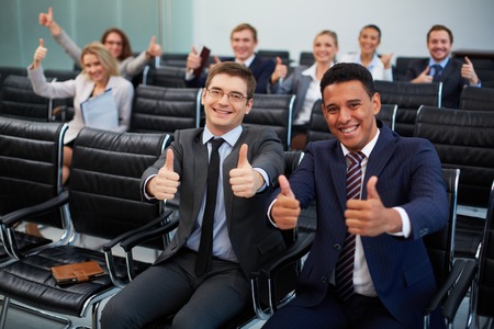 Two happy young businessmen showing thumbs up on background of their colleaguesの写真素材
