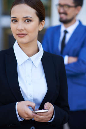 Businesswoman with cellphone looking at camera with elegant man on background