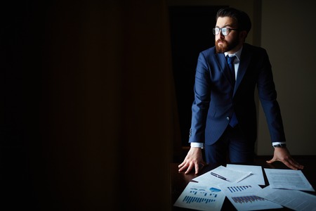 Portrait of elegant businessman in suit and eyeglasses standing by table with papers on itの写真素材