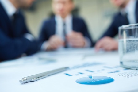 Image of business document and pen at workplace with group of colleagues interacting on background の写真素材