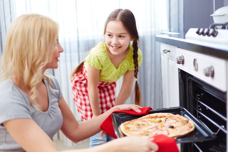 Portrait of young woman taking pizza out of oven with her daughter standing near byの写真素材