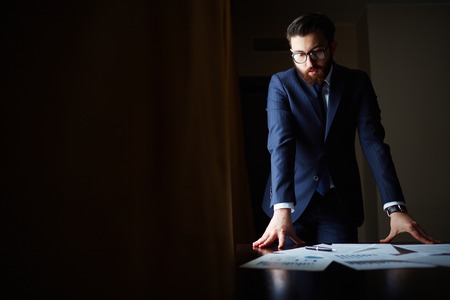 Portrait of elegant businessman in suit and eyeglasses looking at papers on the tableの写真素材