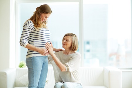 Young woman and her mother looking at pregnant belly and touching itの写真素材