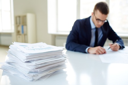 Stack of documents on the desk and male employee working on backgroundの写真素材