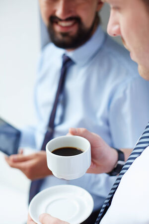 Image of young businessman holding cup of coffee while listening to his colleague at meetingの写真素材