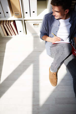 Portrait of young guy in casual clothes sitting on the floor and making notesの写真素材