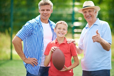Cute boy with rugby ball and his grandfather and father looking at camera outdoorsの写真素材