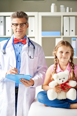 Joyful girl with teddy bear and her doctor looking at camera in hospitalの写真素材