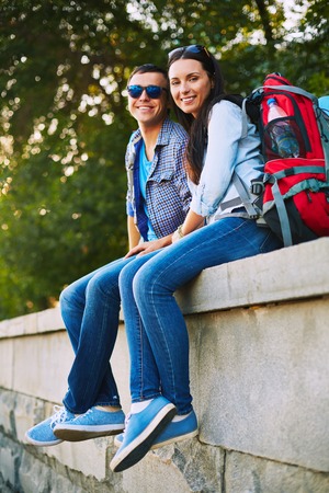 Restful travelers with rucksacks sitting in park and looking at camera
