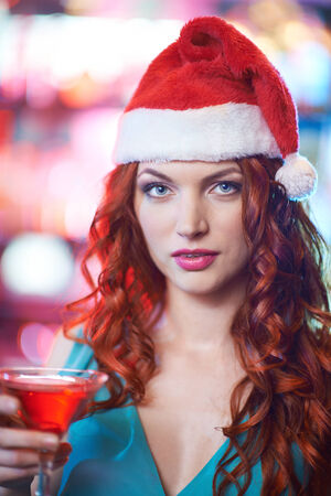 Pretty young girl in Santa cap holding glass of martini and looking at cameraの写真素材