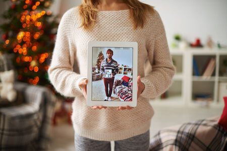 Girl in white knitted sweater showing image of beloved man with her portrait in touchpadの写真素材