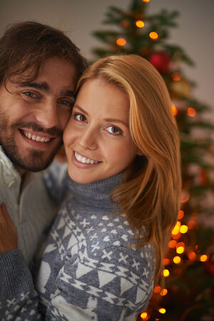 Affectionate couple looking at camera on xmas evening with sparkling firtree on backgroundの写真素材