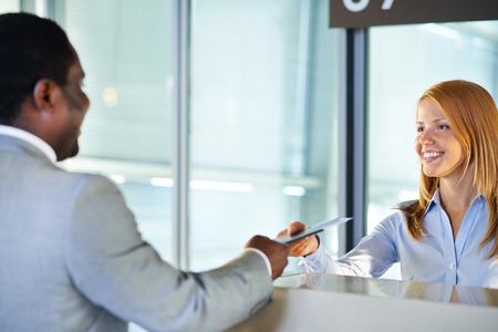Friendly woman giving passport and ticket back to businessman at airport check-in counterの写真素材