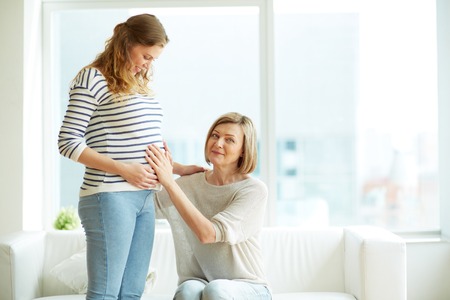 Mature woman touching pregnant belly of her daughter and looking at cameraの写真素材